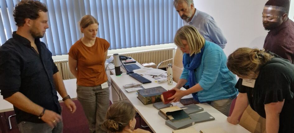 Eine Gruppe des Workshops „(Post-)koloniale Provenienzforschung in Theorie und Praxis“ steht vor einer Sammlung der Staats- und Universitätsbibliothek Hamburg und diskutiert über Provenienzforschung zu Manuskripten.