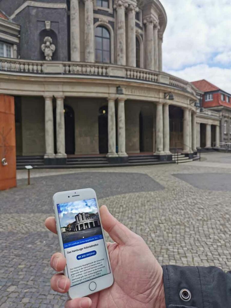 Im Vordergrund ist eine Hand zu sehen, die ein iPhone hält. Auf dem Display ist die App "Koloniale Orte" zu sehen. Die App zeigt Informationen zum "Hamburger Kolonialinstitut".