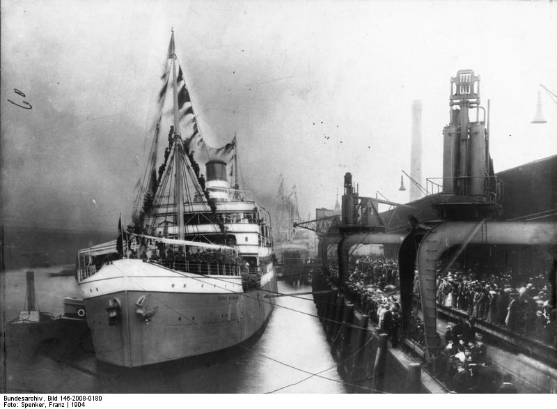 Historisches Foto in schwarz-weiß. Zu sehen ist ein Dampfer im Baakenhafen liegt. Der Dampfer ist eng besetzt mit Personen, die durch die Bildbeschreibung als deutsche Kolonialsoldaten zu identifizieren sind. Auch der Kai ist voller Menschen. 