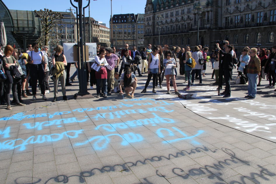 Das Foto zeigt den Rathausmarkt. Auf den Boden wurden Texte in verschiedenen Farben (schwarz, weiß, blau) gesprayt. Um das Geschrieben stehen mehrere Menschen.