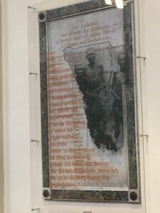 Kommentierte Gedenktafel. Im Hintergrund die marmorne Gedenktafel zum Gedenken an gefallene deutsche Soldaten im Krieg gegen Herero und Nama. Im Vordergrund: Plexiglastafel mit Zitat und Bild aus dem von 2005.