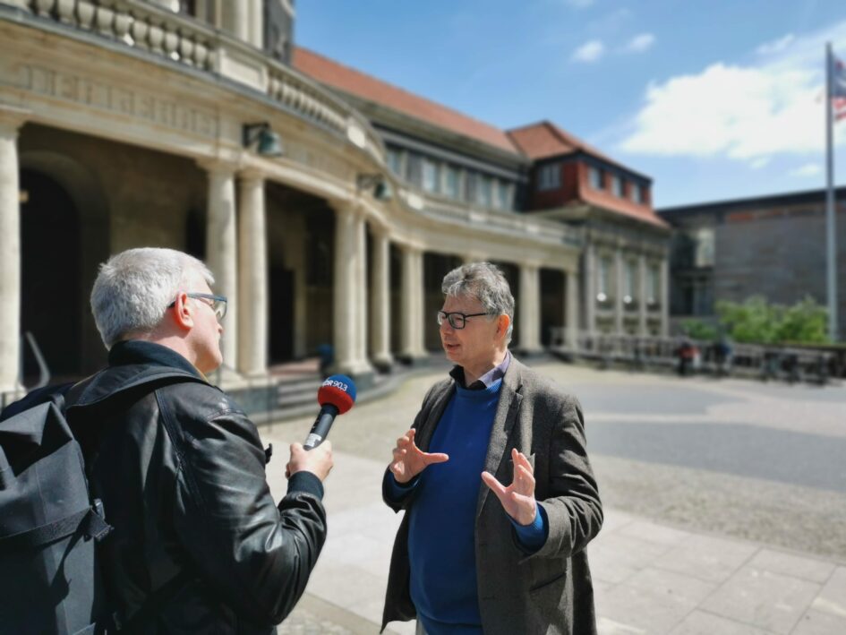 Das Foto zeigt Jürgen Zimmerer im Gespräch mit dem Journalisten Peter Helling. Im Hintergrund ist das Hauptgebäude der Universität Hamburg zu sehen.