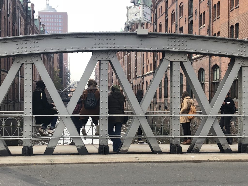 Das Foto zeigt den Blick von einer Straße/Brücke auf die Speicherstadt. Zwischen roten Backsteinfassaden ist ein Kanal zu sehen. Auf der Brücke stehen hinter einer Metallkonstruktion der Brücke mehrere menschen, die auf den Kanal blicken.