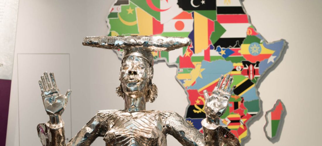 Das Foto zeigt die Skulptur "Looking for Grace" von Sokari Douglas Camp. Im Vordergrund ist eine Frau aus silbernen Metall zu sehen, die die Hände in die Höhe hebt. Im Hintergrund ist der afrikanische Kontinent mit den Flaggen aderafrikanischen Ländern in ihren jeweiligen Staatsgrenzen zu sehen.